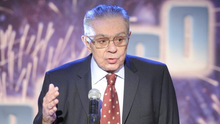 Chico Anysio de terno e gravata fala ao microfone com mão levantada durante apresentação em palco iluminado