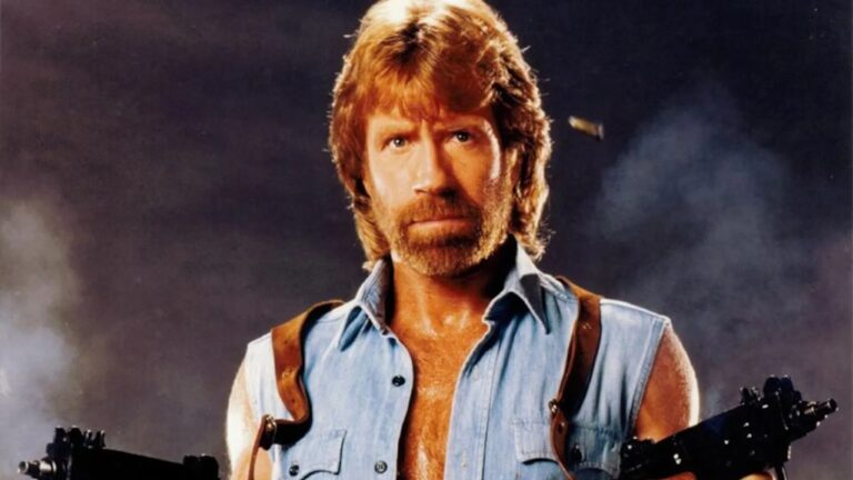Chuck Norris, um homem com barba e cabelo castanho, segura armas, usando camisa sem mangas e coldres, com expressão séria diante de fundo escuro com fumaça.