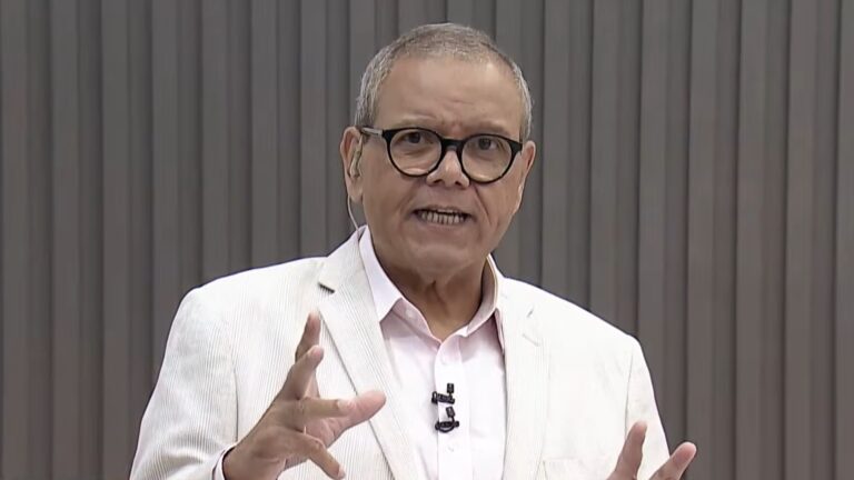 Ciro Bezerra, apresentador do SBT, usa blazer claro e óculos enquanto fala diretamente à câmera em estúdio com fundo neutro, fazendo gesto com as mãos.