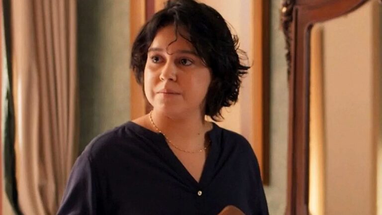 Uma mulher de cabelo curto e escuro está em um ambiente interno, olhando para o lado com expressão séria ou atenta. Ela veste uma blusa escura de manga longa e usa um colar delicado. Ao fundo, há cortinas e um espelho com moldura de madeira, sugerindo um ambiente doméstico. A iluminação é suave e quente, destacando seu rosto e criando um clima íntimo na cena em Três Graças.