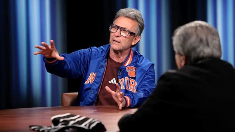 Homem de jaqueta azul do New York Knicks gesticula durante entrevista em programa de TV diante de entrevistador