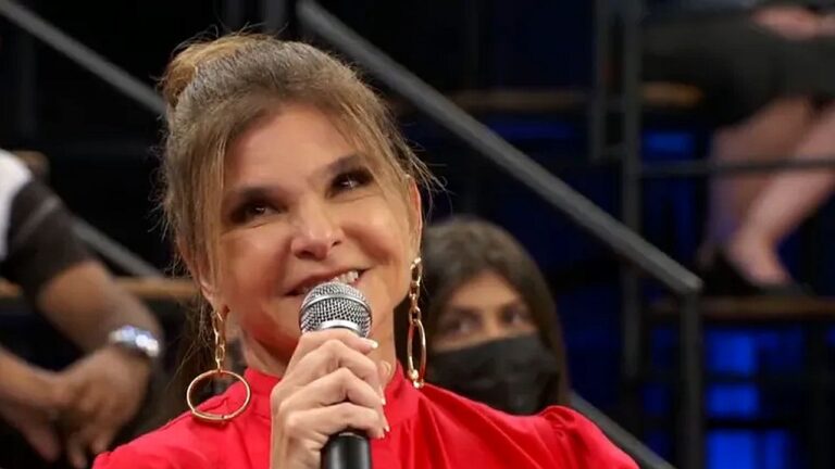 Mulher segura microfone e sorri durante participação em programa de auditório com plateia ao fundo