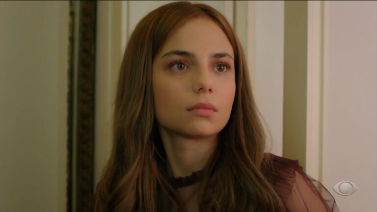 Personagem jovem, com expressão séria, olha para o lado em uma cena de Cruel Istambul, dentro de um ambiente interno com paredes claras; no canto da tela aparece o logotipo da Band.