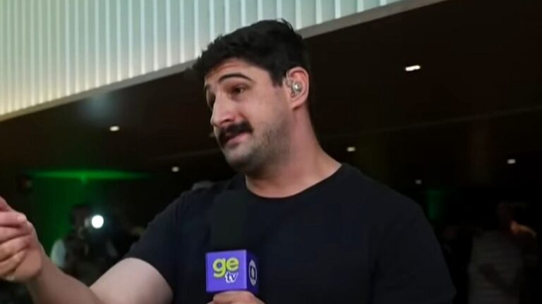 Um homem está sendo entrevistado em um ambiente interno, segurando ou falando próximo a um microfone com o logotipo “ge tv”. Ele usa uma camiseta preta, tem cabelo curto e bigode, além de um fone de ouvido no ouvido direito. Seu corpo está levemente virado para o lado enquanto gesticula com a mão, sugerindo que está explicando algo. Ao fundo, há pessoas e iluminação artificial, indicando um local movimentado, possivelmente um evento ou área de imprensa.