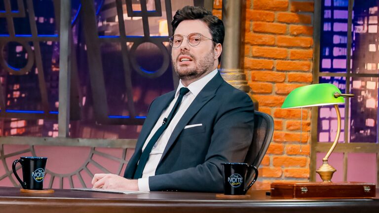 Danilo Gentili, vestindo um terno escuro, camisa branca e gravata verde, está sentado atrás de uma mesa no estúdio do talk show The Noite, usando óculos redondos. Sobre a mesa há canecas pretas com o nome do programa e uma luminária verde, com parede de tijolos e cenário urbano iluminado ao fundo.