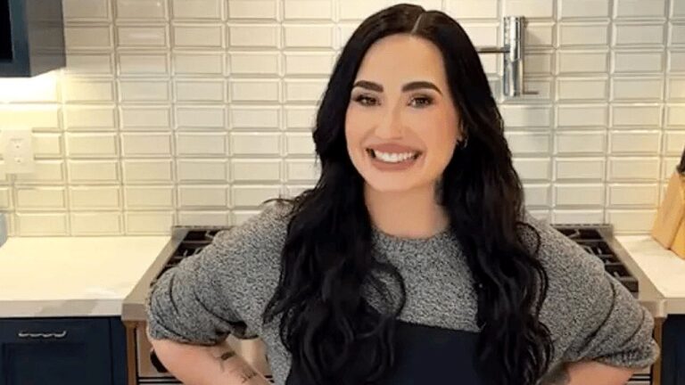 Uma mulher está em uma cozinha moderna, sorrindo diretamente para a câmera. Ela tem cabelos longos e escuros, usa uma blusa de manga longa cinza e um avental preto. Está com as mãos apoiadas na cintura, em uma postura confiante e descontraída. Ao fundo, há um fogão, bancada clara e uma parede com azulejos brancos em padrão geométrico, sugerindo um ambiente organizado e bem iluminado, típico de gravações culinárias.