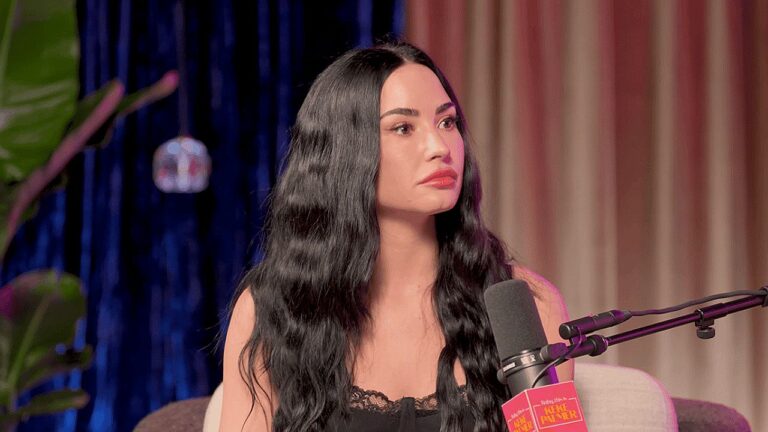 Mulher de cabelos longos e escuros participa de entrevista em podcast falando ao microfone em estúdio com iluminação colorida ao fundo