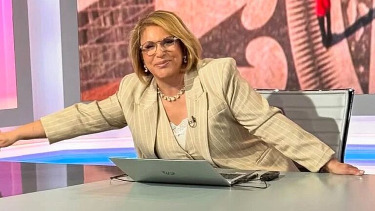 Uma mulher está sentada em um estúdio de televisão, atrás de uma mesa com um notebook aberto à sua frente. Ela usa óculos e um conjunto de blazer claro com listras, além de colar e brincos discretos. Está levemente inclinada para o lado, com um braço apoiado na mesa, sorrindo de forma confiante. O fundo é colorido, com elementos gráficos e iluminação típica de programa televisivo, sugerindo um talk show ou programa de entrevistas.