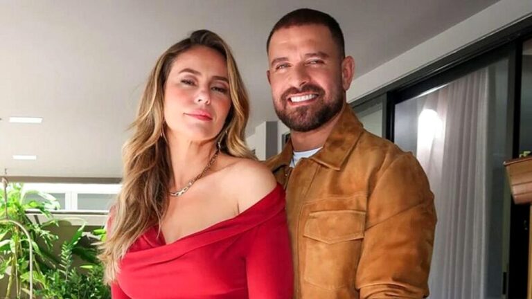 Um homem e uma mulher posando juntos em um ambiente interno; ela usa um vestido vermelho de ombro a ombro e ele veste uma jaqueta marrom, ambos olhando para a câmera.