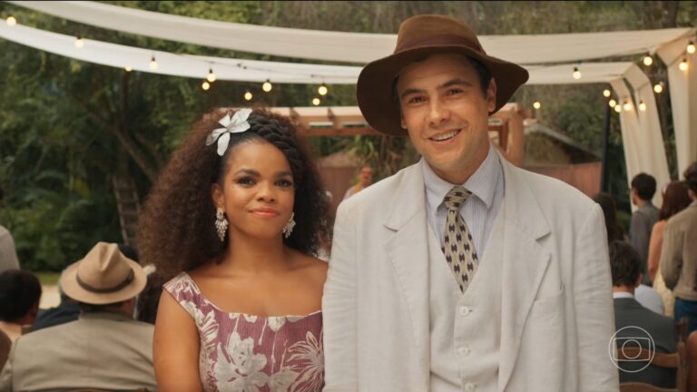 Dita e Candinho, sorridentes, estão lado a lado em uma cerimônia ao ar livre decorada com luzes e tecidos; o homem usa terno claro e chapéu, enquanto a mulher usa vestido estampado e acessório no cabelo, com convidados ao fundo e logotipo da Globo no canto da tela. A imagem é uma cena do último capítulo da novela Êta Mundo Melhor!.