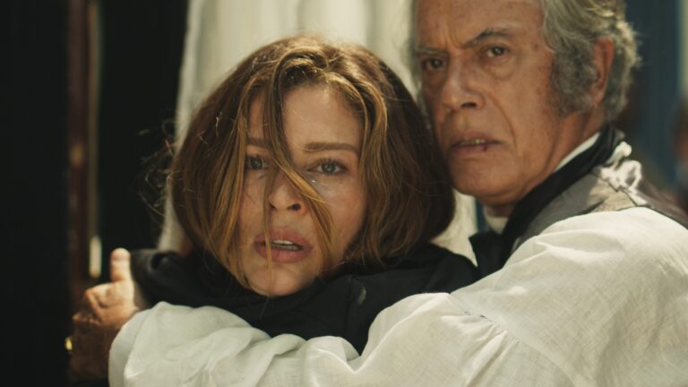 Em Dona Beja, idoso segura uma mulher pela frente enquanto ambos olham assustados para algo fora de cena. A mulher tem os cabelos parcialmente sobre o rosto e expressão tensa, enquanto o homem, vestido com roupas de época, a segura.