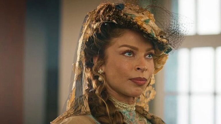 Grazi Massafera em Dona Beja aparece em um ambiente interno, usando roupas de época elegantes, incluindo um vestido detalhado e um chapéu ornamentado com véu. Seu cabelo está estilizado de forma elaborada, com tranças e cachos, e ela usa brincos delicados. A expressão é séria e composta, olhando levemente para o lado. A iluminação suave e o figurino sugerem uma cena ambientada em período histórico ou produção dramática.