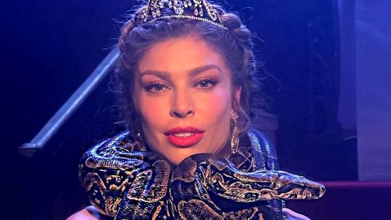 Mulher com tiara e maquiagem marcante aparece em cena de palco com iluminação azul enquanto uma cobra grande está enrolada em seus ombros e pescoço.