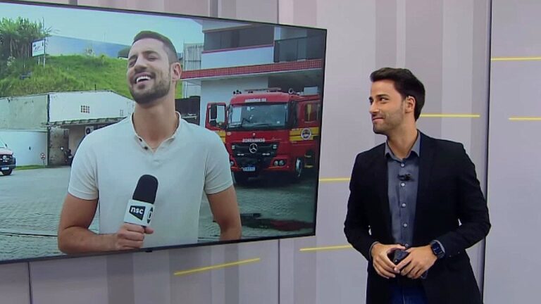 Apresentador observa telão com repórter ao vivo em frente a quartel dos bombeiros durante transmissão de telejornal