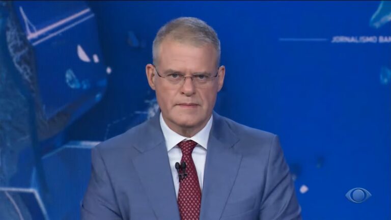 Eduardo Oinegue usa terno azul e gravata vermelha enquanto olha diretamente para a câmera no estúdio do Jornal da Band, com fundo azul e gráficos digitais; no canto da tela aparece o logotipo da Band.