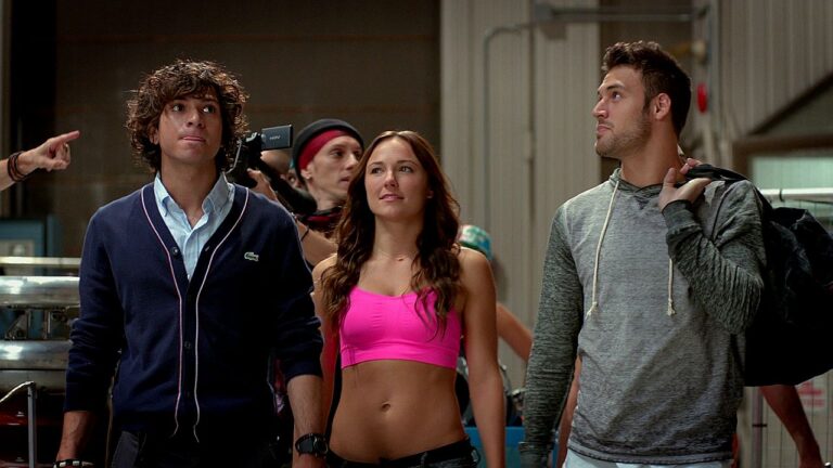 Três jovens caminham lado a lado em um ambiente interno que lembra um estúdio ou local de ensaio. O homem à esquerda tem cabelo cacheado e usa um casaco escuro com camisa por baixo, olhando para cima com expressão curiosa. A mulher no centro veste um top rosa e também observa algo acima, com expressão atenta. À direita, outro homem usa um moletom cinza e carrega uma mochila no ombro, olhando na mesma direção. Ao fundo, há outras pessoas e equipamentos, sugerindo um cenário de produção ou bastidores.