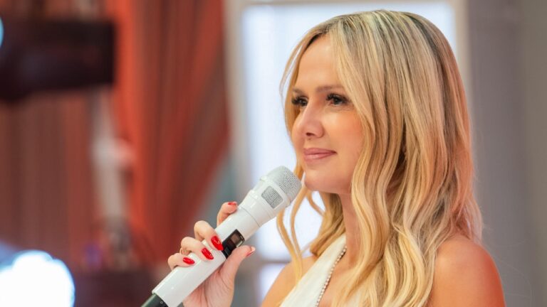 Apresentadora Eliana sorri de perfil, segura um microfone branco próximo ao rosto, possui unhas pintadas de vermelho e veste roupa clara, fundo desfocado