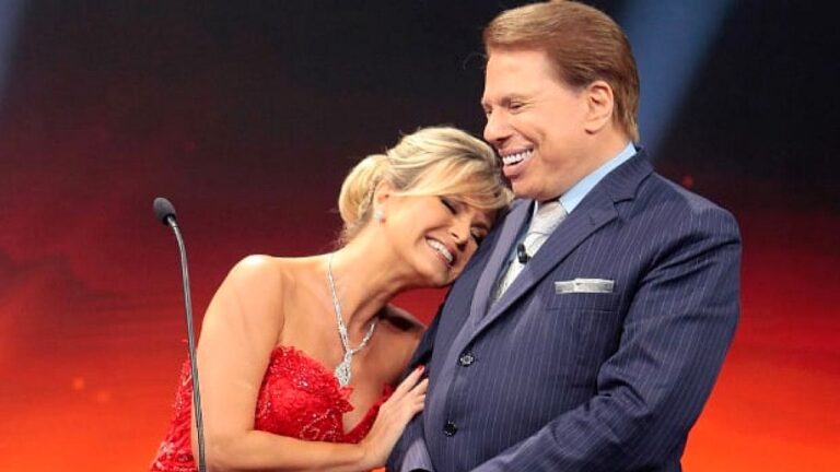 Homem de terno sorri enquanto mulher de vestido vermelho apoia a cabeça em seu ombro durante momento afetuoso em palco de programa de televisão