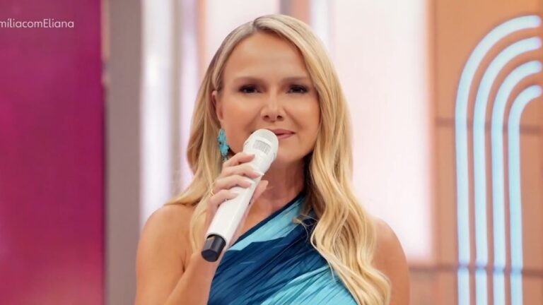 Eliana Michaelichen segura microfone e fala no palco de programa de TV com cenário colorido ao fundo