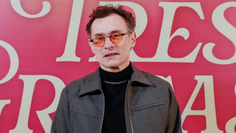 Enrique Diaz usa óculos com lentes alaranjadas e jaqueta escura diante de painel vermelho no lançamento da novela Três Graças