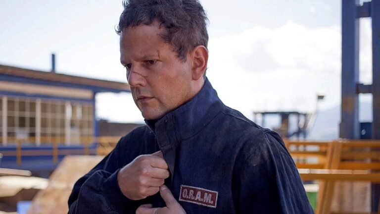 Homem com uniforme escrito Q R A M ajusta gola do casaco em área industrial ao ar livre