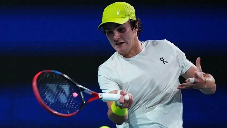 Tenista com boné verde rebate bola com raquete durante partida em quadra dura em jogo profissional de tênis