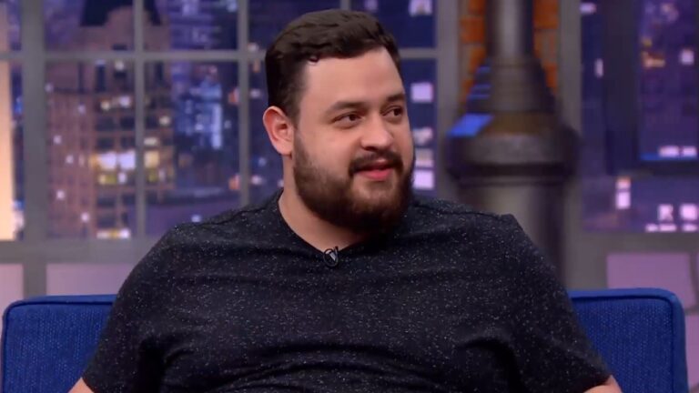 Estevam Nabote sorri levemente enquanto olha para o lado, veste camiseta preta mesclada e está sentado em um sofá azul no cenário do programa The Noite