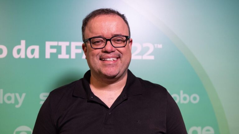Narrador esportivo Everaldo Marques sorri, usa óculos de armação preta e camisa polo escura, posando em frente a painel verde com logos da Fifa e Globo
