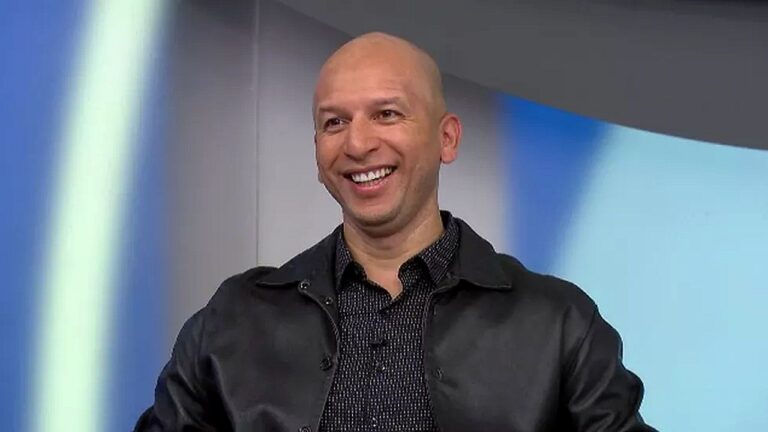 Homem careca sorri durante participação em programa de televisão, sentado em estúdio com fundo iluminado em tons de azul