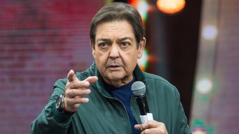 Fausto Silva aponta para frente enquanto segura microfone, veste jaqueta verde escura sobre blusa azul, usa relógio de pulso no palco do Faustão na Band
