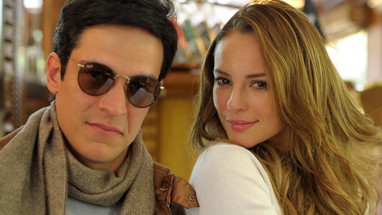 Mateus Solano como Félix usa óculos escuros e cachecol, ao lado de Paolla Oliveira como Paloma, que sorri com cabelos loiros soltos, em cena da novela Amor à Vida