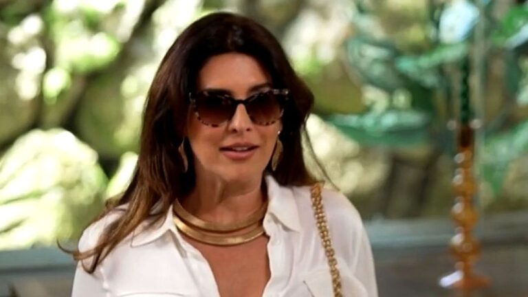 Uma mulher está ao ar livre, usando óculos escuros grandes e uma camisa branca. Ela tem cabelos longos e soltos e usa acessórios dourados, como colar e brincos. Uma bolsa com alça de corrente aparece sobre o ombro. O fundo é desfocado, com elementos de jardim e uma fonte, sugerindo um ambiente externo elegante. A expressão dela é neutra, como se estivesse em uma conversa ou observando alguém.