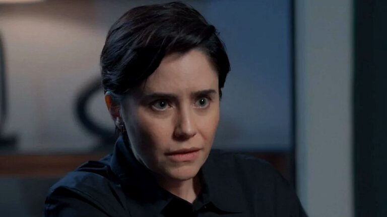 Mulher de cabelo curto e expressão tensa encara alguém fora de quadro durante conversa em ambiente interno, com iluminação suave e fundo desfocado que sugere escritório ou sala reservada.