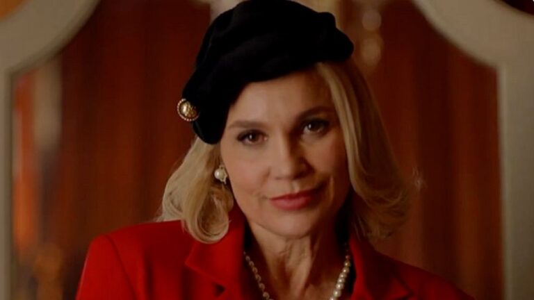 Mulher de blazer vermelho e boina preta encara a câmera em cena de novela ambientada em interior elegante