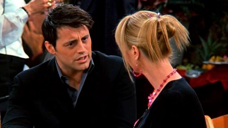 Homem e mulher conversam em cena de série de TV em ambiente interno com outras pessoas ao fundo