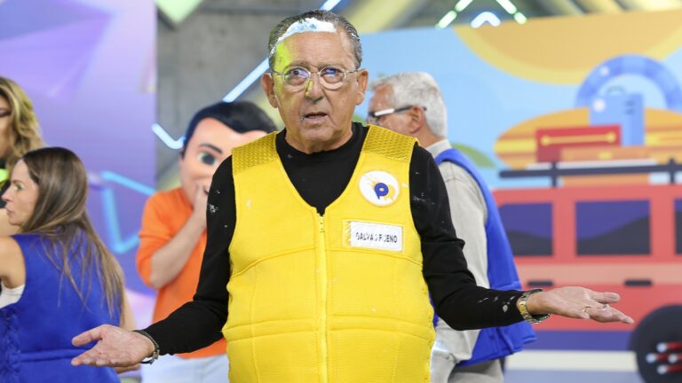 Galvão Bueno, um homem idoso usando óculos e colete amarelo com um crachá, está em um estúdio do SBT, com manchas de torta no cabelo e na roupa, olhando para a frente com expressão surpresa e braços abertos em gesto de dúvida. Ao fundo, outras pessoas participam da cena.