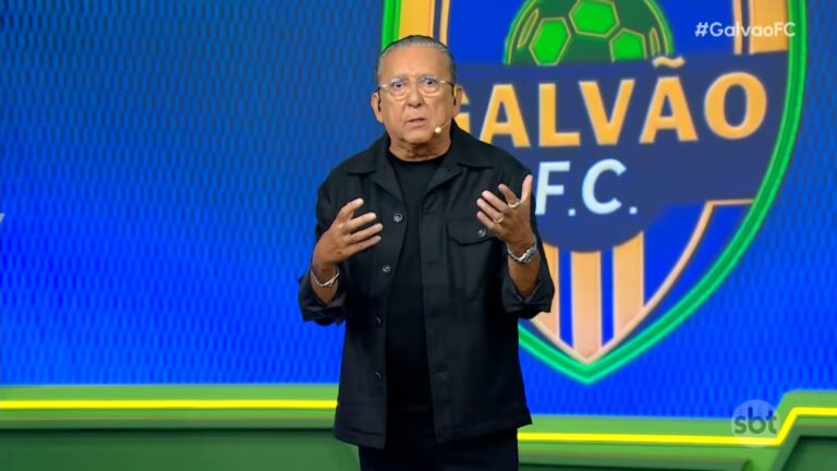 Galvão Bueno, usando roupa preta, fala em estúdio com fundo azul e verde, gesticulando com as mãos. Atrás dele há um grande escudo com o nome do programa e um desenho de bola de futebol. No canto da tela aparece a hashtag do programa Galvão FC, do SBT.