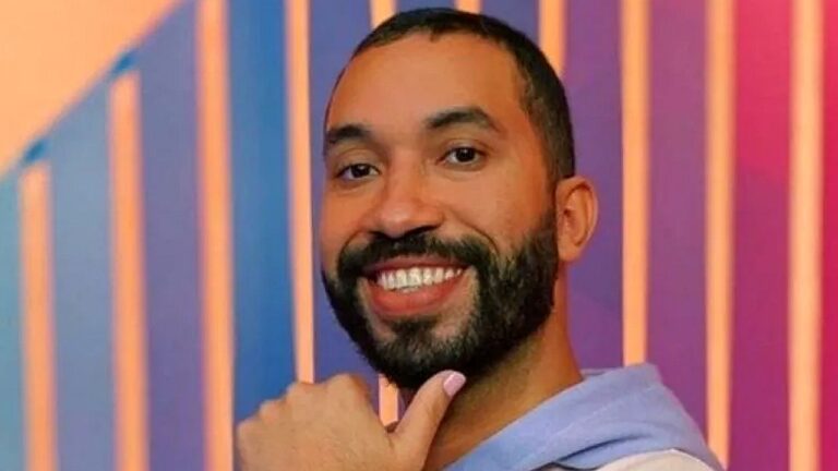 Homem sorri para a câmera com mão no queixo em cenário colorido de estúdio com luzes ao fundo