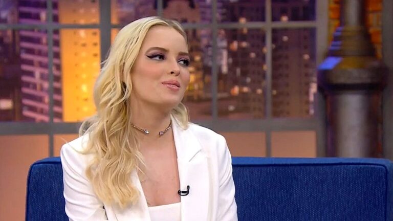 Mulher loira participa de entrevista em programa de talk show sentada em sofá de estúdio com cenário urbano iluminado ao fundo