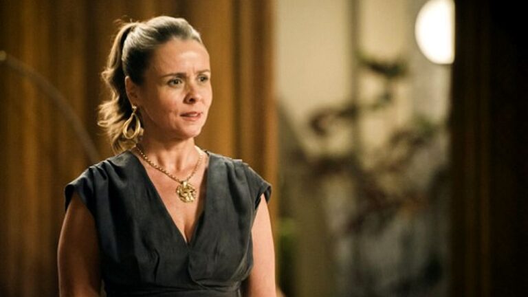 Mulher de vestido preto e rabo de cavalo fala com expressão séria em cena de novela em ambiente interno elegante