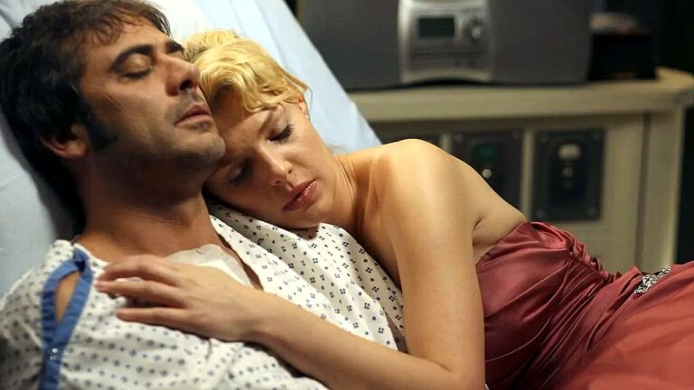 Um homem está deitado em uma cama de hospital usando roupa hospitalar, enquanto uma mulher de vestido vermelho se deita ao lado dele e apoia a cabeça em seu peito, em um momento íntimo e emocional.