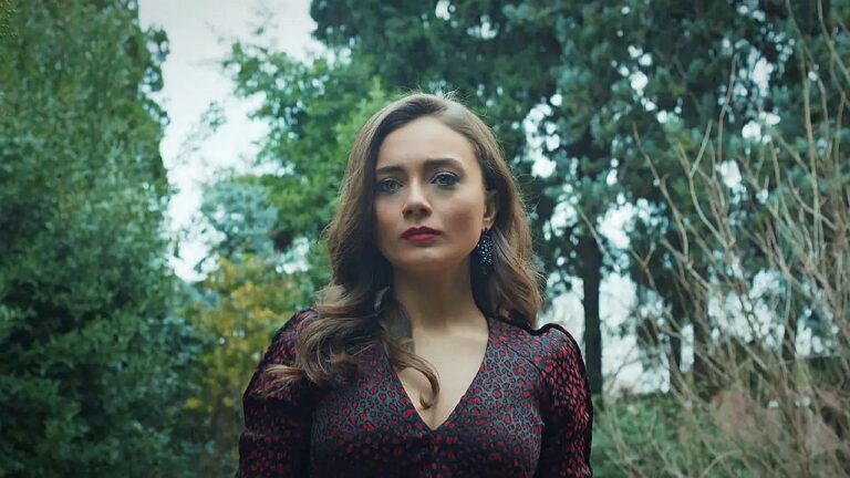 Mulher em um ambiente de floresta, usando um vestido escuro com detalhes vermelhos e brincos, olhando à frente com expressão séria.