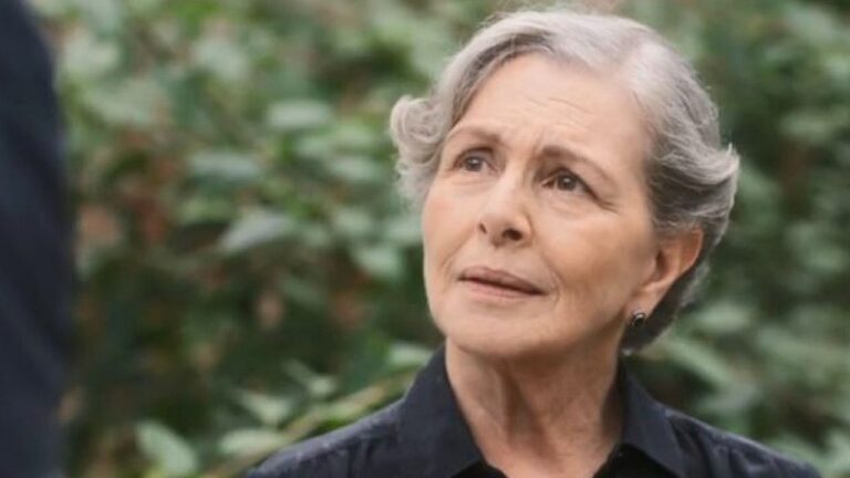 Uma mulher idosa aparece em primeiro plano, com cabelos curtos grisalhos cuidadosamente penteados. Ela veste uma camisa escura e olha para alguém fora de cena com expressão séria e levemente preocupada. O fundo é desfocado, com vegetação verde, indicando um ambiente externo. A iluminação natural destaca as linhas do rosto e reforça o tom emocional da cena.