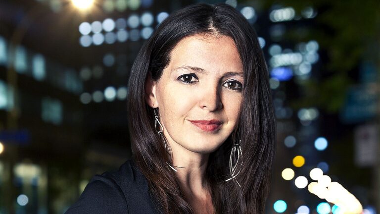 Mulher de cabelo escuro usa brincos longos e encara a câmera com cidade iluminada ao fundo à noite