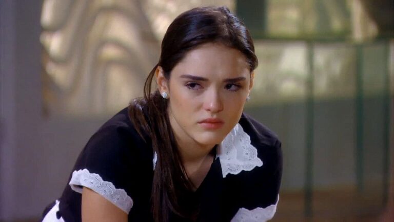 Jovem com uniforme de empregada doméstica demonstra tristeza em cena de novela dentro de casa