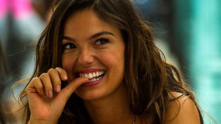 Mulher jovem sorri amplamente enquanto morde levemente o dedo, em momento descontraído com fundo desfocado de ambiente urbano ou comercial