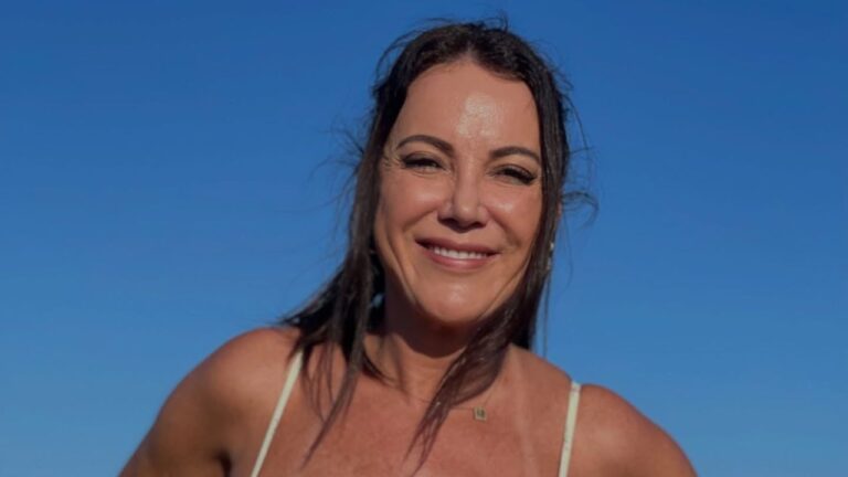 Ivy Mena sorri para a câmera com os cabelos levemente bagunçados pelo vento, usando blusa de alças finas clara sob um céu azul limpo e ensolarado