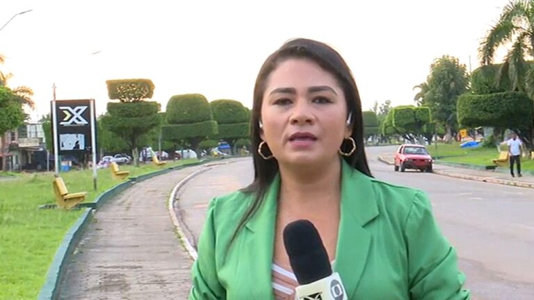 Uma mulher aparece em pé à beira de uma rua, segurando um microfone, como em uma reportagem externa. Ela usa blazer verde e brincos, com expressão séria e focada, olhando diretamente para a câmera. Ao fundo, há uma via com carros passando, bancos ao longo da calçada e árvores podadas em formato geométrico, sugerindo um ambiente urbano organizado.