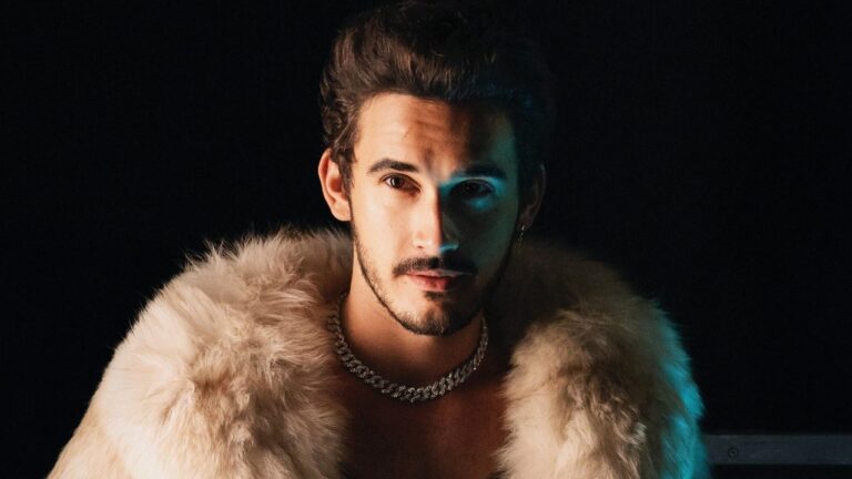 Filipe Bragança como João Raul posa com casaco de pele e colar em foto promocional da novela Coração Acelerado com fundo escuro