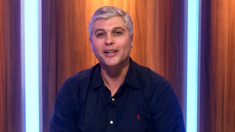 Joel Datena sorri durante participação em estúdio de TV com painel de madeira e luzes verticais ao fundo